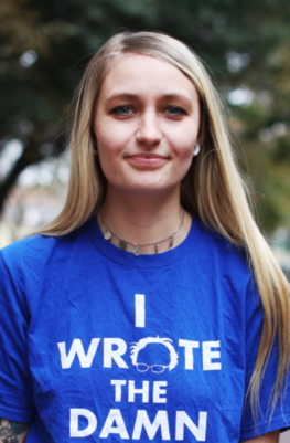 A white woman with long blonde hair and a necklace wearing a blue 'I Wrote The Damn Bill' t-shirt